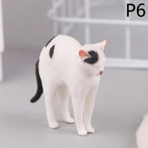 May include: A small, white cat figurine with black spots and a black tail. The cat has a black spot on its head and pink accents on its ears and nose. The figurine is standing on a gray surface.