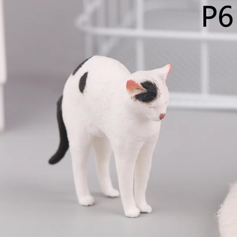 May include: A small, white cat figurine with black spots and a black tail. The cat has a black spot on its head and pink accents on its ears and nose. The figurine is standing on a gray surface.