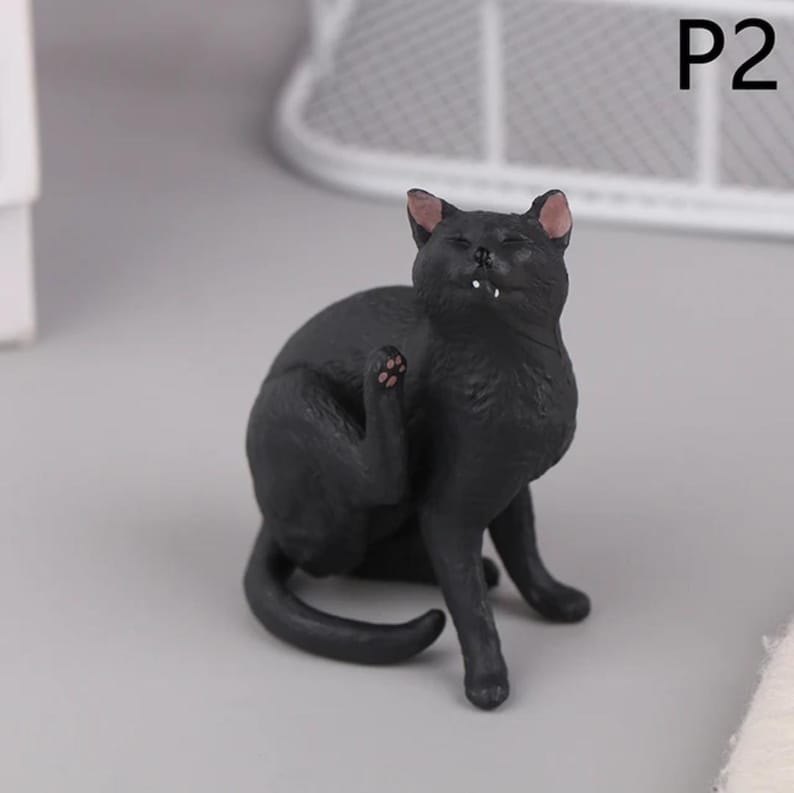 May include: A black cat figurine in a seated position, scratching its ear. The cat has pink inner ears and paw pads. The figurine is on a gray surface, with the text "P2" in the upper right corner.