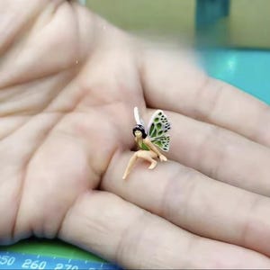 May include: A miniature fairy figurine with butterfly wings. The fairy is wearing a green outfit and has black hair. The wings are white with green spots. The figurine is small and detailed.