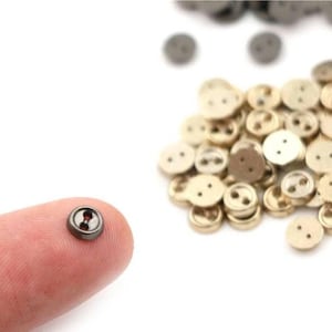 May include: A selection of small, round buttons in shades of gold and grey. One button is held on a finger, showing its dark grey rim and red centre. The buttons have two or four holes for sewing and are scattered on a white background.