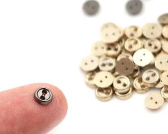 Può includere: Una collezione di piccoli bottoni rotondi in varie tonalità di oro e grigio. Un bottone è tenuto su un dito, mostrando il suo bordo grigio scuro e il centro rosso. I bottoni hanno due o quattro fori per cucire e sono sparsi su uno sfondo bianco.