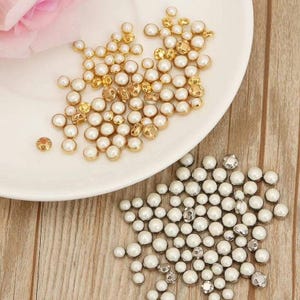 May include: A top-down view displays a white plate with gold and white pearl beads. Below, silver and white pearl beads are scattered. These beads, ideal for crafts and jewellery, come in various sizes and shapes.