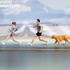 May include: Three miniature figures running on a path. A woman in a grey shirt and blue shorts, a man in a grey tank top and black shorts, and a golden brown dog.