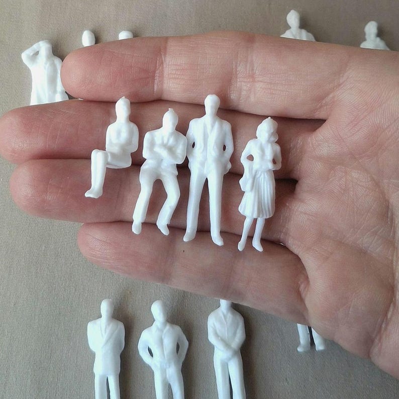 May include: Miniature white plastic figures in various poses, including standing, sitting, and with arms crossed. These small figurines are likely used for architectural models or dioramas. The figures are held in a hand.