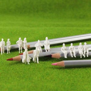 May include: Small white figurines of people are positioned on grey pencils with red and pink tips, against a green backdrop. Some figurines are standing, others are seated, creating a miniature scene.
