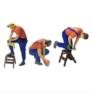 May include: Three miniature figures of construction workers wearing orange and blue overalls. One figure is standing on a small wooden step stool, another is kneeling on the ground, and the third is standing on a step stool while holding a tool.
