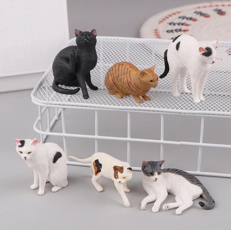 May include: A collection of six cat figurines in various poses and colors. The cats include black, orange tabby, white with black spots, and white with brown and black markings. They are arranged on a white wire shelf.