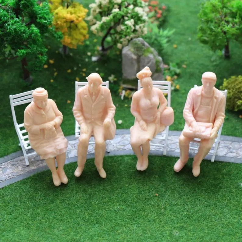 May include: Four miniature plastic figures of people sitting on white chairs. The figures are all wearing different clothes and have different hairstyles. The figures are all in a seated position and are facing forward. The figures are all in a park setting.