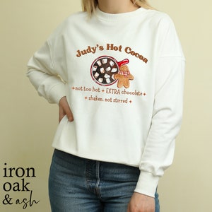 Op de afbeelding: Witte trui met een bruine en rode afbeelding van een mok met warme chocolademelk met marshmallows en een peperkoekman. De tekst "Judy's Hot Cocoa" staat in een bruin lettertype boven de afbeelding. De tekst "not too hot + EXTRA chocolate + shaken. not stirred +" staat in een bruin lettertype onder de afbeelding.
