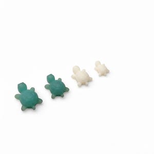 May include: Four small turtle figurines in varying sizes and colors. Two are a translucent teal, and two are off-white. The turtles are arranged diagonally on a white background, showcasing their detailed shell and head features.