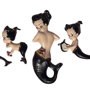May include: Four vintage mermaid figurines. Each mermaid has black hair and a black fishtail. The mermaids have a light skin tone and are in various poses, one holding a mirror, one with a flower, and one playing a harp.