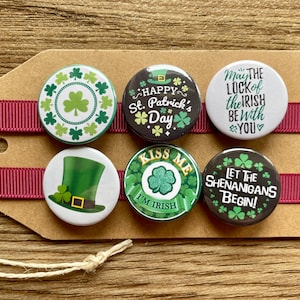 May include: Set of six round black and white buttons with green shamrock designs and St. Patrick's Day themed text. The buttons read "Happy St. Patrick's Day", "Kiss Me I'm Irish", "Let the Shenanigans Begin", "May the Luck of the Irish Be With You", and feature a shamrock design.