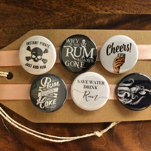 May include: Set of six round black and white buttons with various rum-themed sayings. The buttons are arranged on a brown card with pink ribbon.