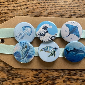 May include: Six blue and white round buttons with different ocean-themed illustrations. The buttons feature a jellyfish, a wave, a manta ray, a seahorse, a sea turtle, and a whale shark.