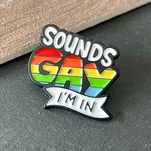 Puede incluir: Pin de esmalte negro con rayas arcoíris y el texto "Sounds Gay I'm In".