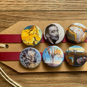 May include: Six round, colorful, printed buttons featuring famous Salvador Dali artwork. The buttons are arranged in two rows of three on a brown card with red ribbon.