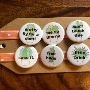 Może przedstawiać: Sześć okrągłych białych guzików z ilustracjami kaktusów i zabawnymi powiedzeniami.  Powiedzenia to "pretty fly for a cacti", "me so thorny", "can't touch this", "succ it.", "free hugs" i "little prick".