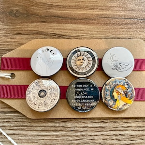 May include: Six round, black and white, pinback buttons with astrological themes. The buttons feature images of the sun, moon, stars, hands, and zodiac symbols. One button has the text "Astrology is a language. If you understand this language, the sky speaks to you."