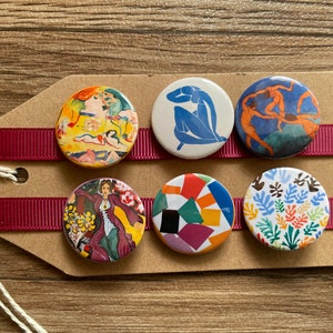 May include: Six round buttons featuring various colourful abstract art designs. The buttons are arranged in two rows of three on a brown card with red ribbon.