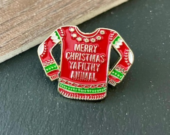 Può includere: Spilla smaltata rossa a forma di maglione natalizio. La spilla presenta il testo bianco "MERRY CHRISTMAS YA FILTHY ANIMAL". Il design del maglione include dettagli verdi e bianchi.