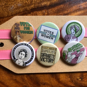May include: A collection of six circular Suffragette badges. The badges display slogans and images, such as "Votes for Women" and "Suffragette Victory." They are presented on a brown tag, secured with pink ribbon.