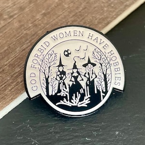 May include: A circular enamel pin in black and white, inscribed with "GOD FORBID WOMEN HAVE HOBBIES". The design shows three witch silhouettes, a full moon, and bare trees.