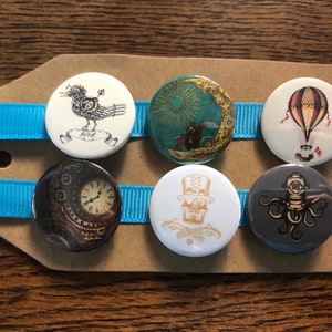 Puede incluir: Seis botones redondos, blancos, negros y dorados con diferentes diseños de temática steampunk. Los diseños incluyen un pájaro, un reloj, una calavera, un globo aerostático, una luna y un pulpo.
