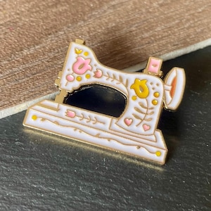 May include: A gold-toned metal enamel pin shaped like a sewing machine. The pin features a white base with decorative floral patterns in pink and yellow, including small heart shapes and vine motifs. The design includes a dark cutout area.