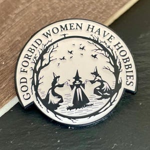 Pode incluir: Pin redondo de esmalte preto e branco com o texto "GOD FORBID WOMEN HAVE HOBBIES". A imagem central mostra três bruxas de mãos dadas, com uma árvore e pássaros ao fundo. Um acessório decorativo.