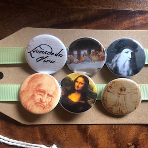 Può includere: Sei bottoni rotondi bianchi con immagini di opere d'arte di Leonardo da Vinci e una firma. I bottoni mostrano immagini dell'Ultima Cena, della Gioconda, dell'Uomo Vitruviano e un ritratto di Leonardo da Vinci. La firma recita "Leonardo da Vinci".