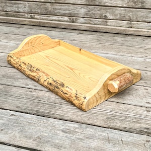 May include: A rectangular wooden serving tray with a natural edge and a handle. The tray is made of light-colored wood with visible grain and a bark edge. The handle is a small, cylindrical piece of wood with a textured surface.