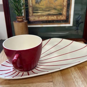 May include: A red and white striped ceramic saucer with a red ceramic teacup on top. The saucer is shaped like a fan.