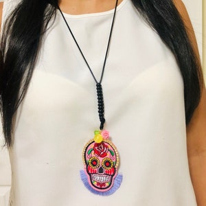 May include: A pink and black sugar skull necklace with a purple tassel. The skull is embroidered with flowers and has gold accents.