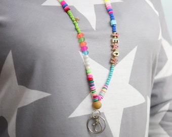 Puede incluir: Un cordón de cuentas de colores con un dije dorado y un llavero. Las cuentas son de varios colores, incluyendo rosa, azul, verde y amarillo. El cordón tiene la palabra "TEACH" escrita en letras doradas.