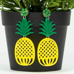 May include: A pair of yellow pineapple earrings with green leaves and gold accents. The earrings are made of plastic and have a cutout design.