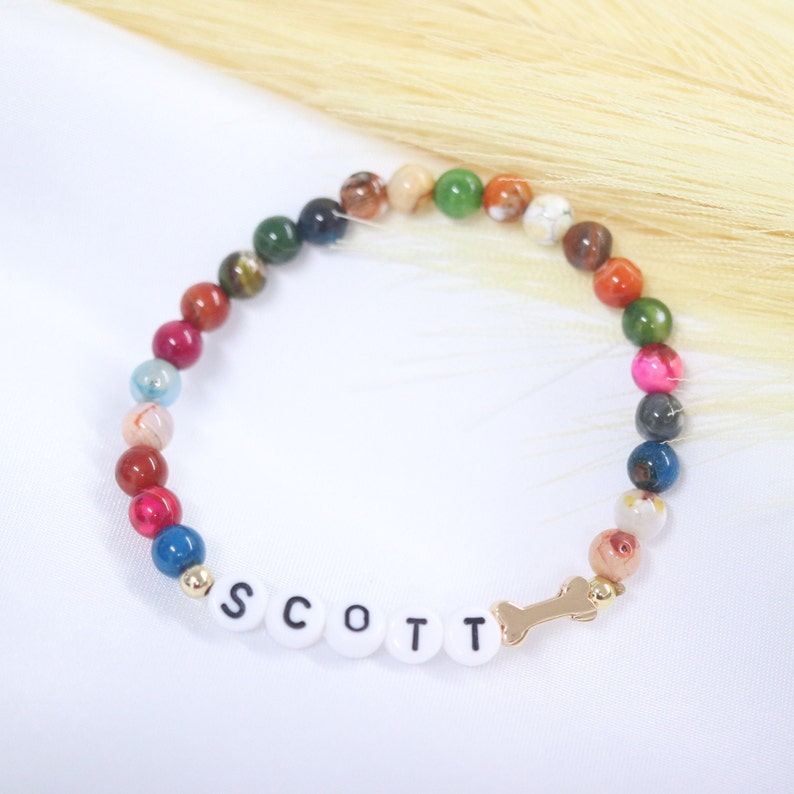 Puede incluir: Un brazalete de cuentas de colores con un dije de hueso dorado y el nombre "SCOTT" escrito en cuentas blancas.