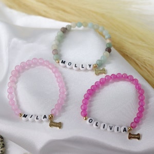 Peut inclure: Trois bracelets de perles aux couleurs rose et vert. Chaque bracelet comporte des perles blanches &eacute;pelant un nom et un charm en forme d'os dor&eacute;. Les breloques indiquent "I <3 MY DOG". Les bracelets sont pr&eacute;sent&eacute;s sur une surface blanche.
