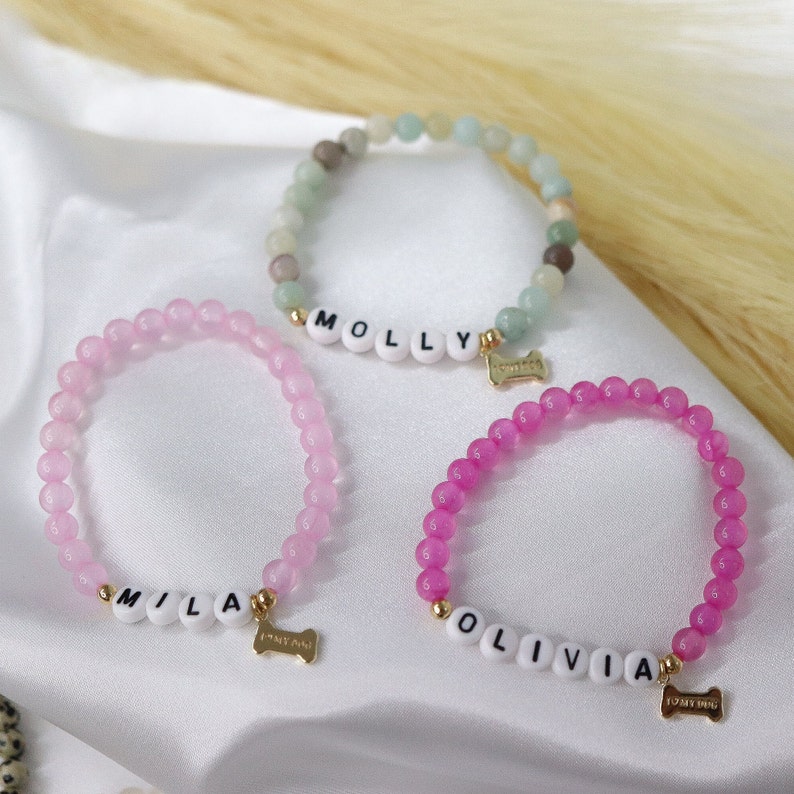 Peut inclure: Trois bracelets de perles aux couleurs rose et vert. Chaque bracelet comporte des perles blanches &eacute;pelant un nom et un charm en forme d'os dor&eacute;. Les breloques indiquent "I <3 MY DOG". Les bracelets sont pr&eacute;sent&eacute;s sur une surface blanche.