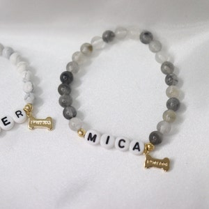 Peut inclure: Deux bracelets de perles avec le nom "MICA" &eacute;pel&eacute; en perles de lettres blanches. Les bracelets pr&eacute;sentent des perles grises et blanches, des accents dor&eacute;s et un charm en forme d'os de chien dor&eacute; avec l'inscription "I MY DOG".