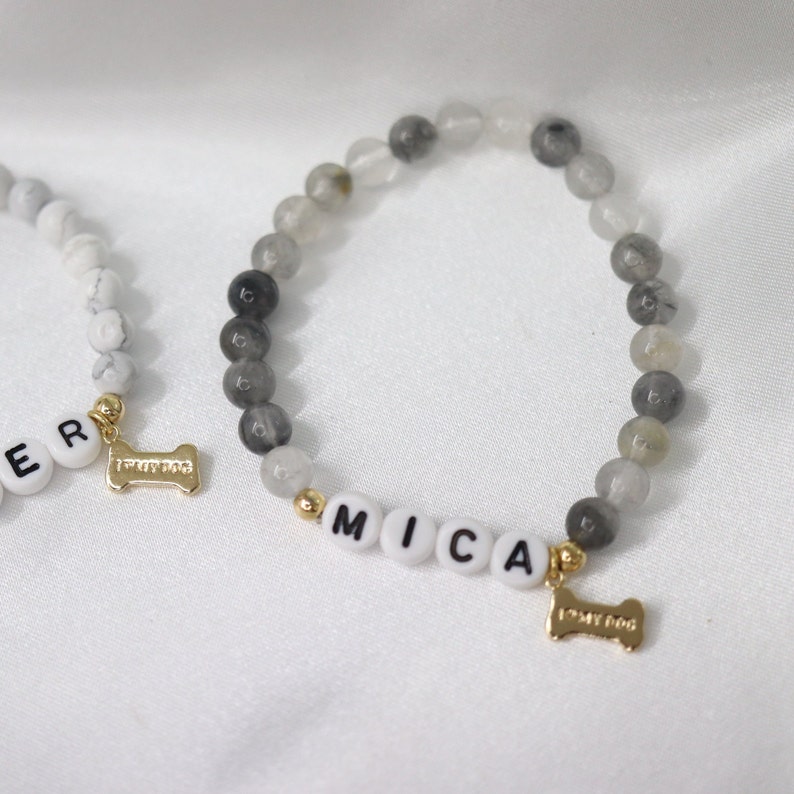 Peut inclure: Deux bracelets de perles avec le nom "MICA" &eacute;pel&eacute; en perles de lettres blanches. Les bracelets pr&eacute;sentent des perles grises et blanches, des accents dor&eacute;s et un charm en forme d'os de chien dor&eacute; avec l'inscription "I MY DOG".