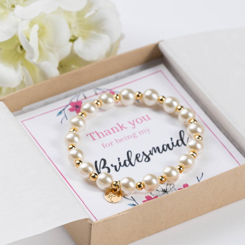 May include: A white pearl bracelet with a gold charm that says "Thank you for being my Bridesmaid". The bracelet is in a small gift box with a floral design.