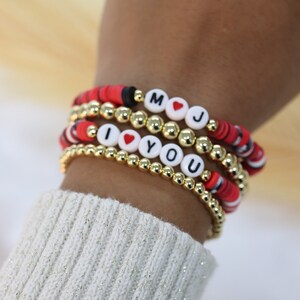 May include: A set of four bracelets with gold beads and white letter beads spelling "I love you" and "MJ". The bracelets are red and black with a striped pattern.