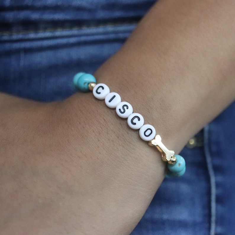 Puede incluir: Un brazalete de cuentas turquesa y doradas con el nombre "CISCO" escrito en letras blancas y un dije de hueso dorado.
