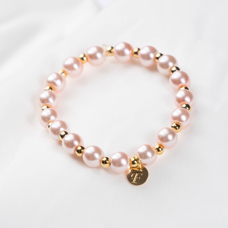 May include: A delicate bracelet made of pink pearls and gold beads. The bracelet has a gold charm with the letter "F" on it.
