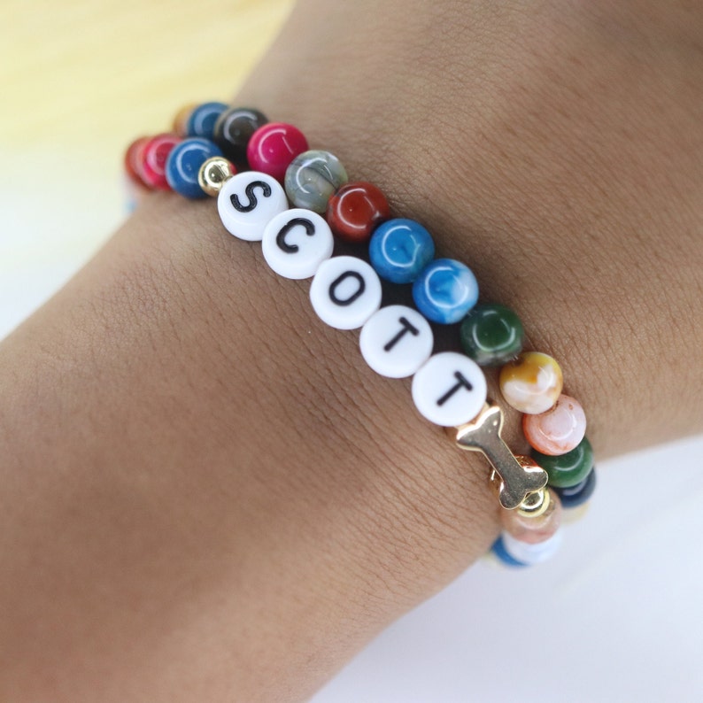 Puede incluir: Un brazalete de cuentas multicolores con un dije de hueso dorado y cuentas de letras blancas que deletrean el nombre "SCOTT".