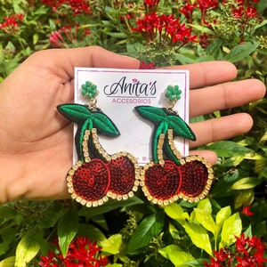 May include: A pair of cherry earrings with red sequins and green felt leaves. The earrings are on a white card with the text "Anita's Accesorios".