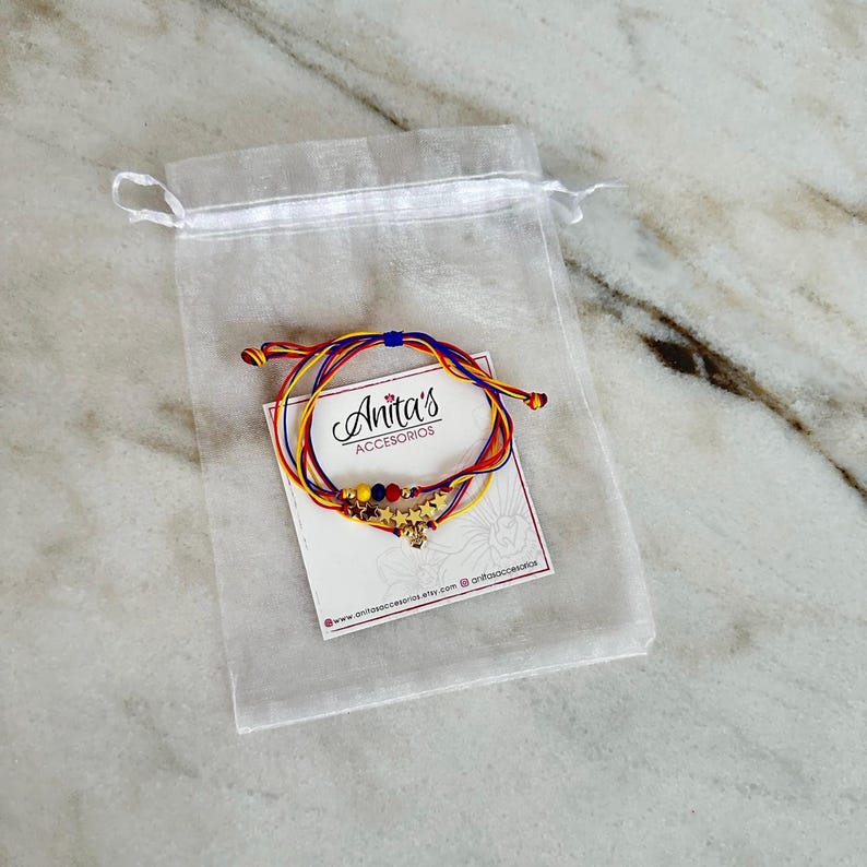 May include: A set of three colorful string bracelets with gold star charms and beads. The bracelets are packaged in a white organza bag with a small card that reads "Anita's Accesorios" and includes the website address.