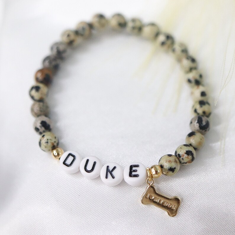 Peut inclure: Un bracelet de perles avec le nom "DUKE" &eacute;pel&eacute; en perles de lettres blanches. Le bracelet comporte des perles de pierre tachet&eacute;es et un charm en forme d'os dor&eacute; avec l'inscription "I LOVE DOG".
