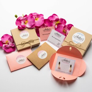 Puede incluir: Un conjunto de cajas de regalo con orqu&iacute;deas rosas y una caja peque&ntilde;a que contiene un par de pendientes de perlas. Las cajas est&aacute;n decoradas con papel marr&oacute;n y cuerda, y cada caja tiene una pegatina con el texto "Anita's Accessories".